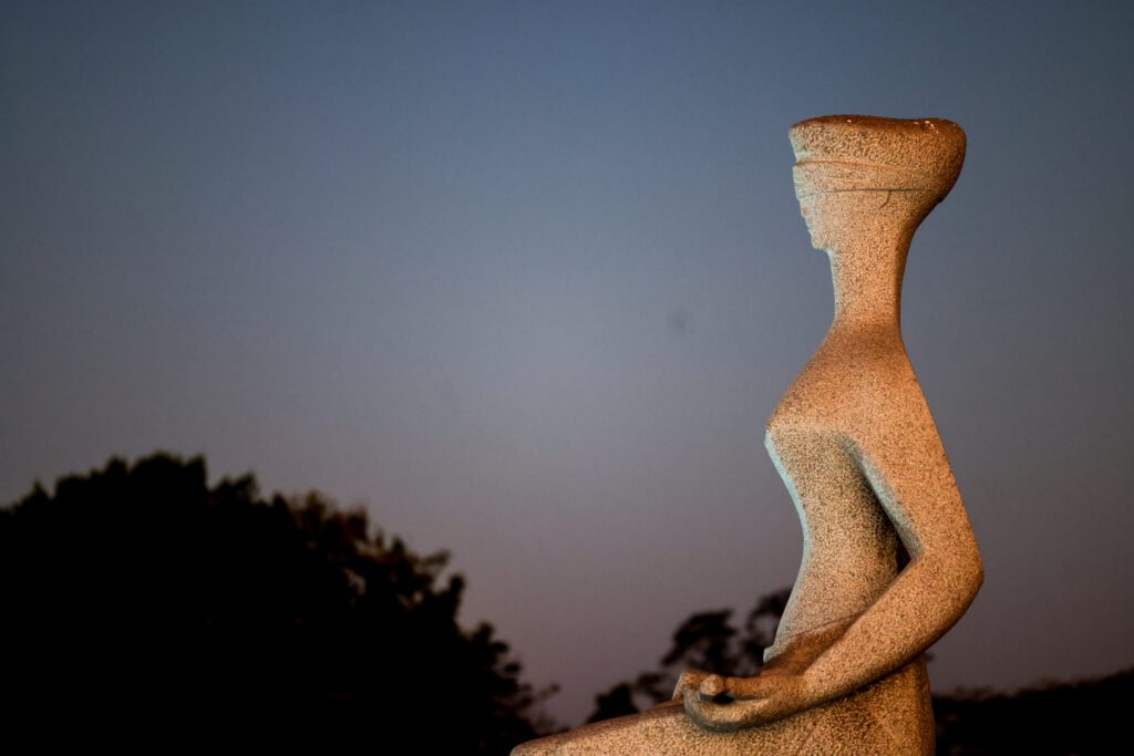 Foto colorida da estátua da justiça em momento de crepúsculo. Ela é vista de lado, ao fundo da imagem aparece a copa de uma árvore e o céu anoitecendo.